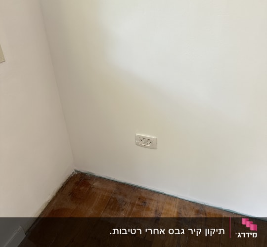 פינת חדר עם קיר לבן חלק, שקע חשמל ורצפת פרקט